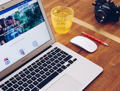 A MacBook Air displaying a webpage about Facebook ads is on a wooden desk. Next to the laptop is a white mouse, a red pencil, a glass filled with yellow plastic stirrers, and a black camera.