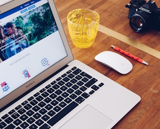 A MacBook Air displaying a webpage about Facebook ads is on a wooden desk. Next to the laptop is a white mouse, a red pencil, a glass filled with yellow plastic stirrers, and a black camera.