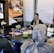 A group of people are gathered in a conference room, engaged in a UX training session. Two women are seated at a table with laptops, participating in the training. A large poster on the wall advertises UX Indonesia and a UX training series. Papers with diagrams are taped to the wall, and various electronic devices and cups are on the table.