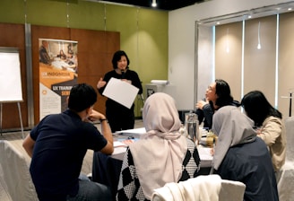 A group of people is engaged in a training session in a conference room setting. A presenter is standing and holding a sheet of paper, addressing the group seated at a round table. The attendees are actively listening, and there's a banner in the background advertising UX training. The room is well-lit, with a flipchart visible at one side.