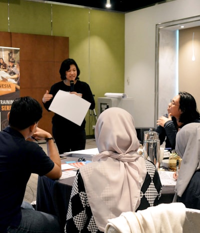 A group of people is engaged in a training session in a conference room setting. A presenter is standing and holding a sheet of paper, addressing the group seated at a round table. The attendees are actively listening, and there's a banner in the background advertising UX training. The room is well-lit, with a flipchart visible at one side.