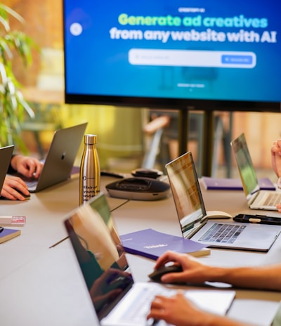 A conference room setting with several laptops on a large table, each being used by a person. A large screen displays a blue interface with the text 'Generate ad creatives from any website with AI'. A stainless steel water bottle and a conference phone are also visible on the table.