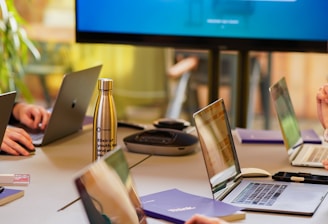 A conference room setting with several laptops on a large table, each being used by a person. A large screen displays a blue interface with the text 'Generate ad creatives from any website with AI'. A stainless steel water bottle and a conference phone are also visible on the table.